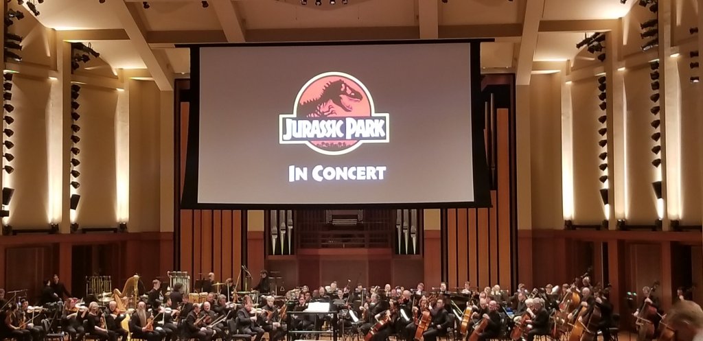 Projection screen above the Seattle Symphony, with the logo/title of Jurassic Park in Concert on it.