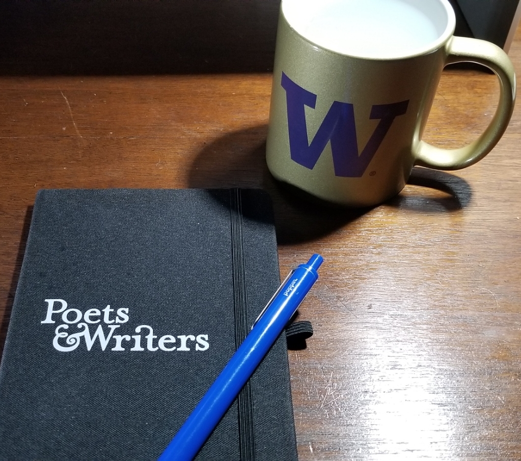 Black journal with the words "Poets & Writers" on the cover in white. Elastic band keeps the book closed. Atop the journal, a blue pen.

To the upper right of that, a gold colored cup with a purple W on it for the University of Washington. The cup is filled with water.