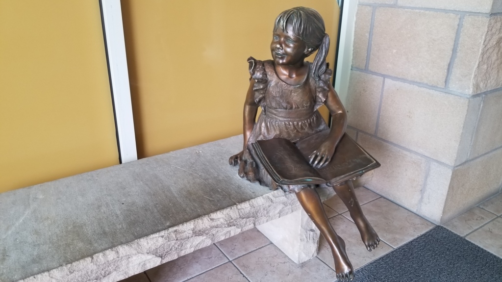 Bronze statue of a pig-tailed girl on a concrete bench. She has a hand resting on an open book, looking out towards the entrance.
