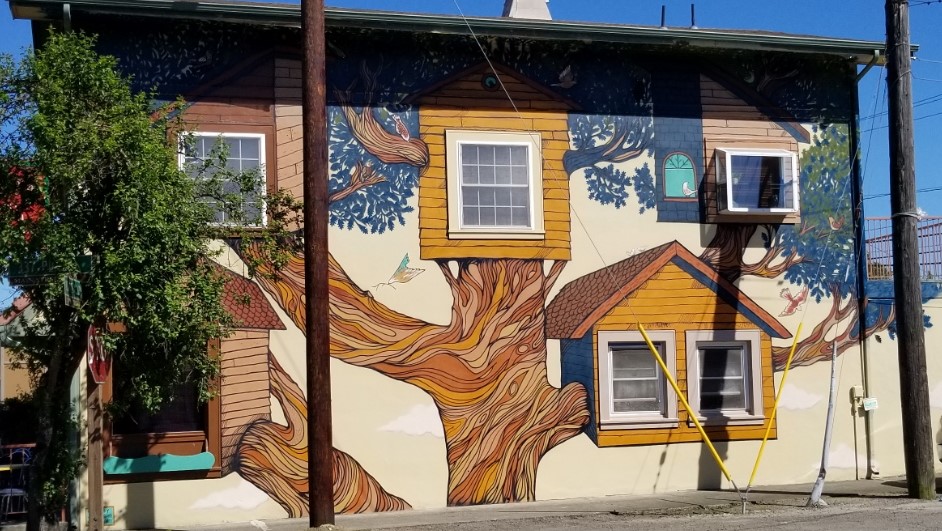 A mural painted on the side of a restaurant. Image is of a large tree, with the restaurant windows and AC unit being used as windows of smaller treehouses.