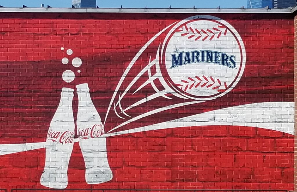 Red background with white swipe of the Coca Cola logo, with two Coca Cola bottle silhouettes on the left, with bubble above them, and swoosh lines beside a Mariners logoed baseball on the right. 