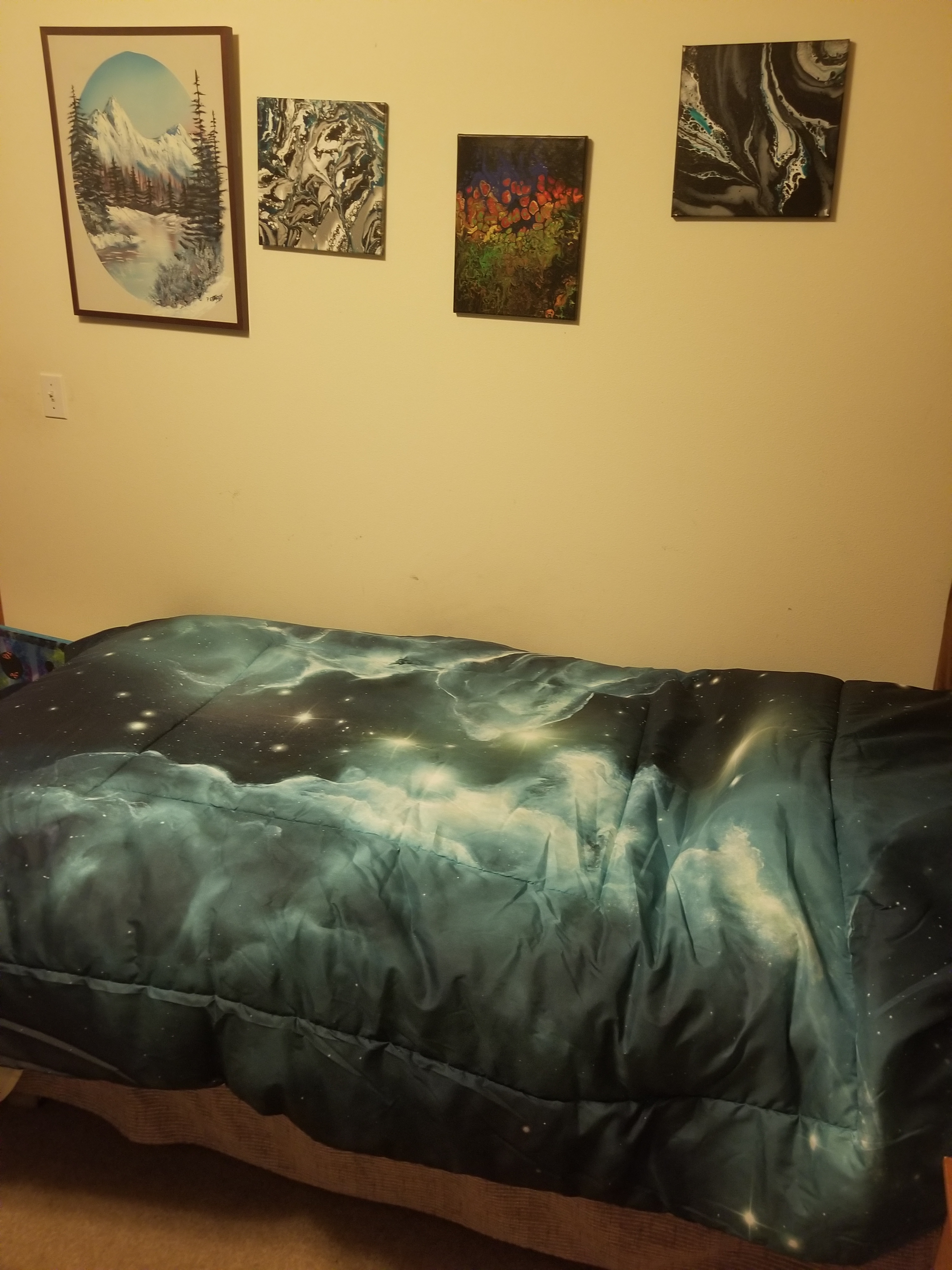 A twin- sized comforter of the stars, with a painting of a mountain, and three abstract paintings on the wall above the bed. 