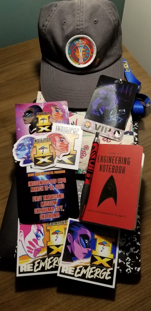 A collection of stickers from IndigiPop, a miniature Star Trek notebook, a ballcap with Jeffrey Veregge's Space Needle design
